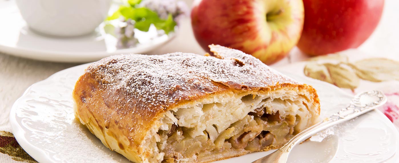 Fetta di strudel di mele con zucchero a velo su briciole, piatto bianco e mele rosse fresche sullo sfondo